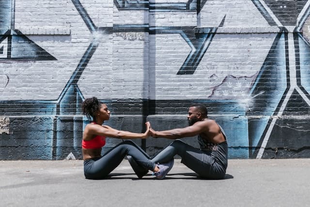 Foto de um casal de pessoas pretas sentadas em uma quadra de rua, de frente um para o outro, encontando seus pés e mãos. A foto ilustra o artigo O que é Calistenia do site Me reinvento.